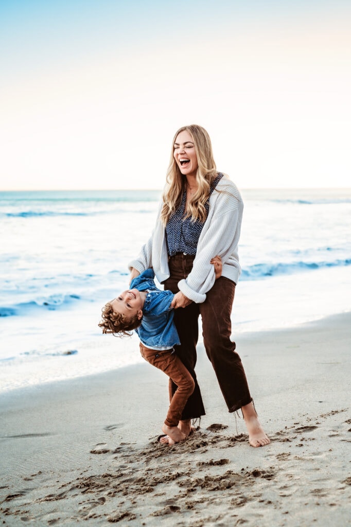 Family Photographer, a mother hangs on to her toddler playfully as he leans backward on the beach sand