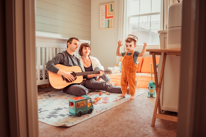 Family Photographer, a man and woman sit in a nursery with the guitar, their toddler dances
