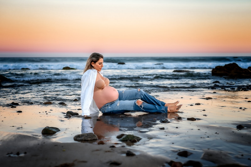 Maternity Photographer, a pregnant woman sits in the sand near the tides at sunset
